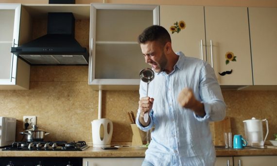 Attractive young funny man dancing and singing with ladle while cooking breakfast in the kitchen at home in the morining