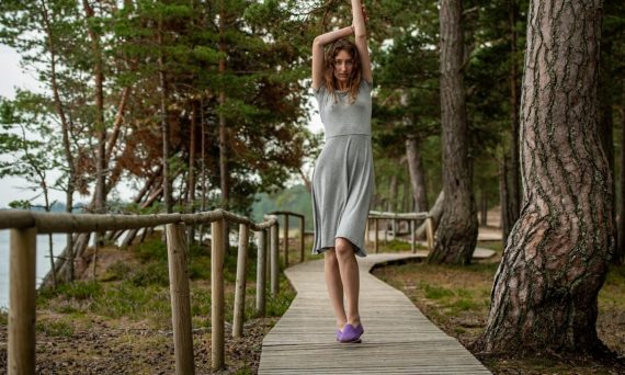 A slim dark-haired woman in a gray dress walking along a wooden boardwalk by the sea, surrounded by coastal forest.