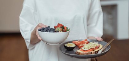 Person holding a bowl of fresh berries and a plate with avocado toast, displaying a healthy breakfast spread.